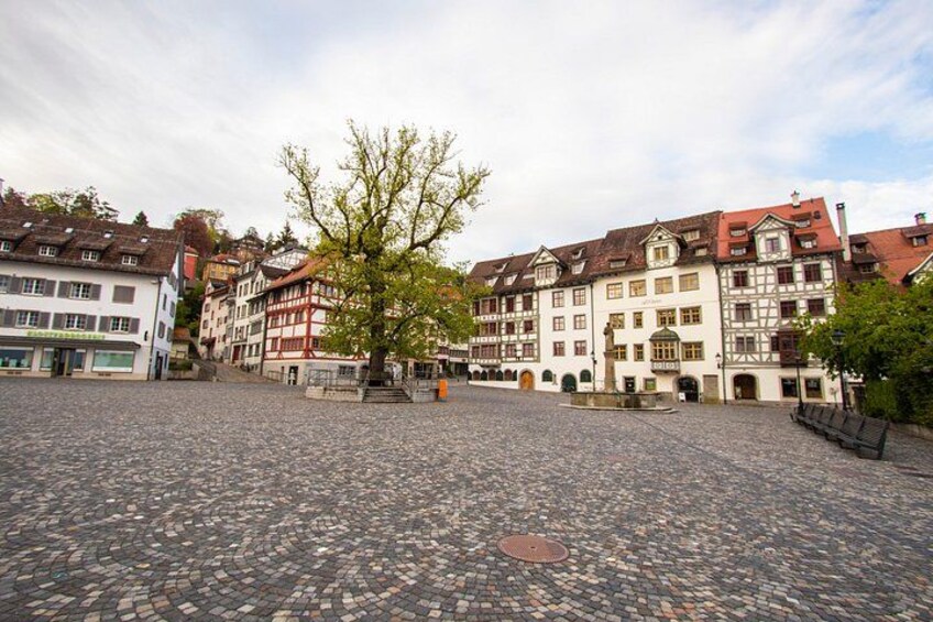 A cobblestone square with historic buildings, a tree, and a bench. 2