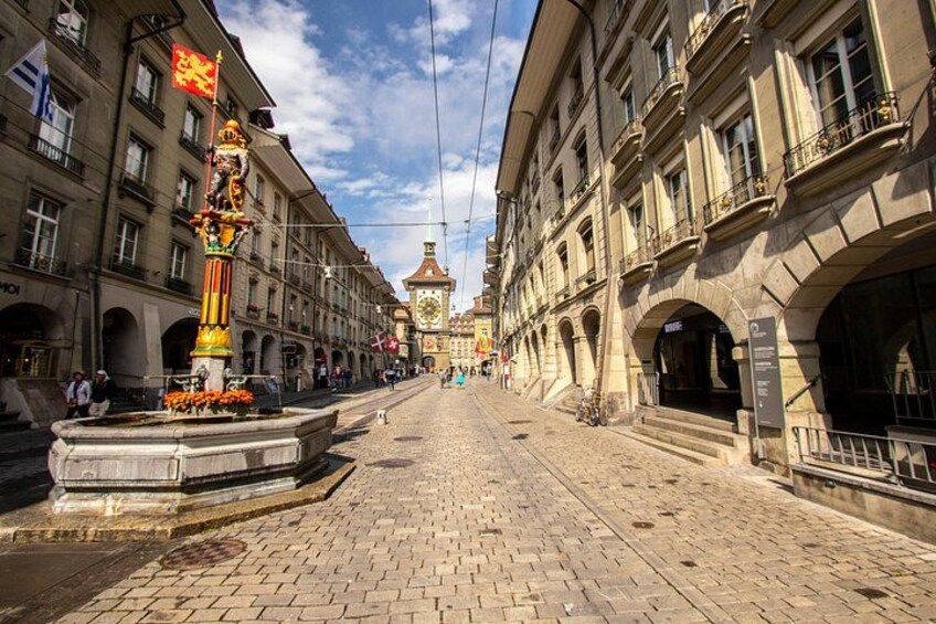 A cobblestone street lined with historic buildings, a prominent fountain, and a clock tower in the distance. 2