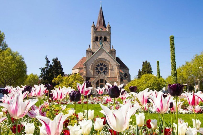 Een kerk met een klokkentoren, omgeven door een tuin vol tulpen en andere bloemen. 1