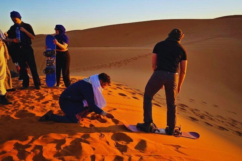 Un gruppo di persone nel deserto, tra dune di sabbia: una persona è inginocchiata, un'altra in piedi con una snowboard. 5