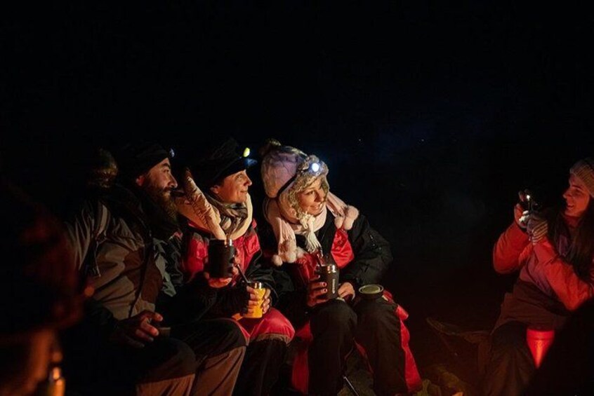 Un groupe de personnes assises à l’extérieur, de nuit, dont certaines tiennent des verres à la main. 2