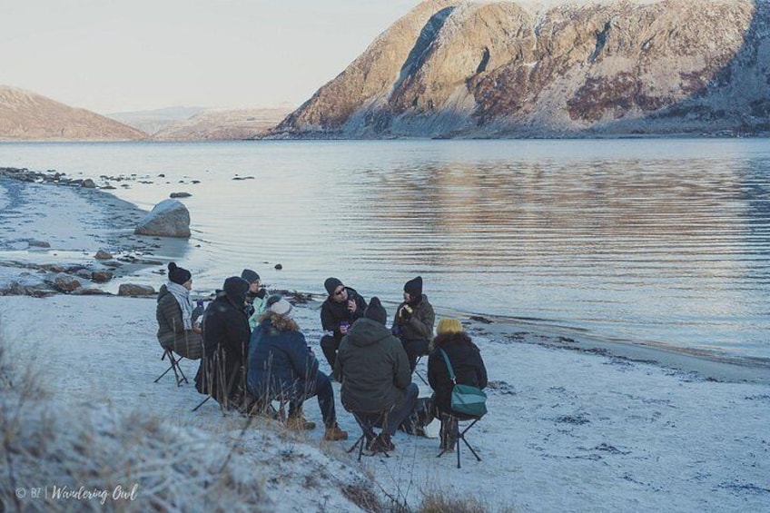 Eine Gruppe von Menschen sitzt auf Stühlen am Ufer eines zugefrorenen Sees, im Hintergrund Berge. 4