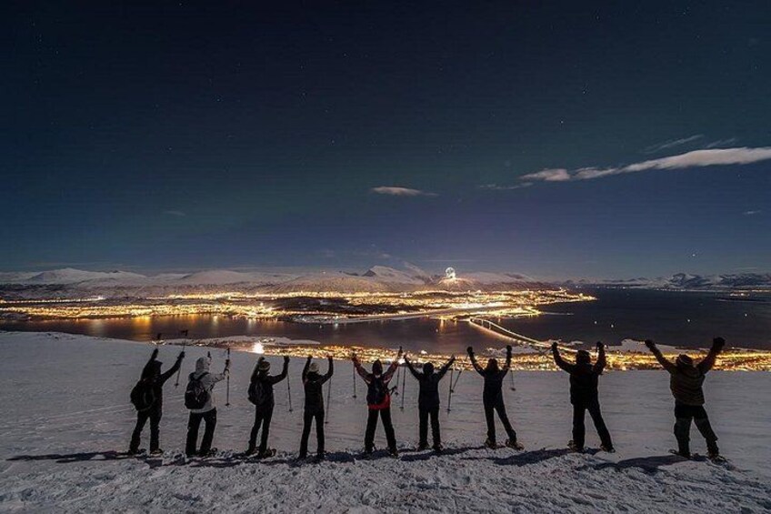Een groep mensen staat op een besneeuwde ondergrond, met armen omhoog, kijkend naar een stadsbeeld met lichtjes en bergen op de achtergrond. 4