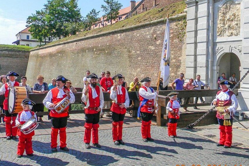 En grupp människor i traditionella dräkter uppträder utomhus, med en historisk byggnad i bakgrunden. 3