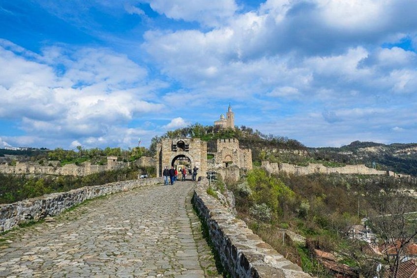 Un sendero de piedra que lleva a un castillo histórico con una torre de campanas, rodeado de colinas y árboles. 4