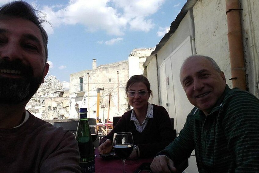 Three people sitting at a table with drinks, a bottle, and a glass, in an outdoor setting with buildings in the background. 1