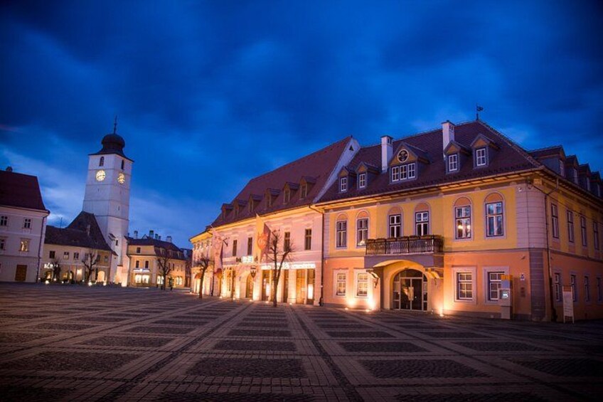 A historic square with a prominent clock tower and illuminated buildings at twilight. 3