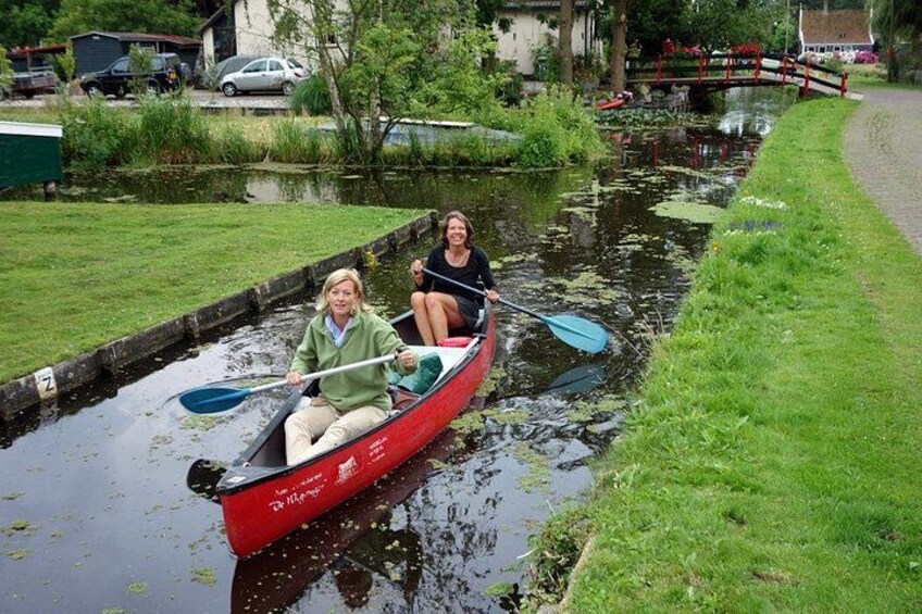 Due persone su una stretta chiatta rossa in un canale, circondate dal verde e con un piccolo ponte sullo sfondo. 5
