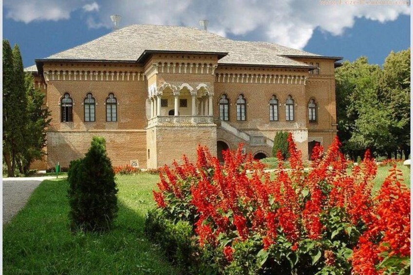 A large brick building with arched windows and a central entrance, surrounded by a well-maintained garden with red flowers. 1