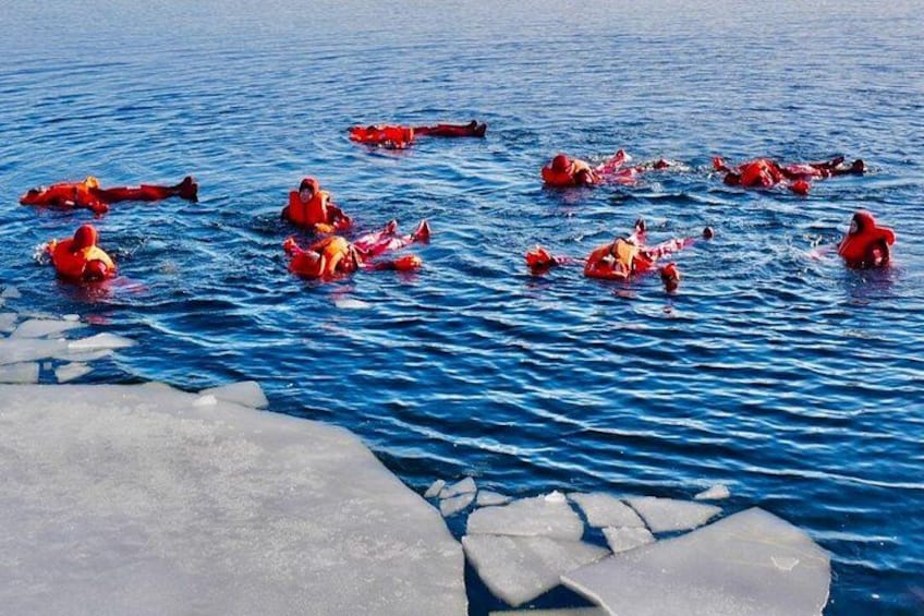 People in red life jackets floating on a body of water with icebergs in the foreground. 1