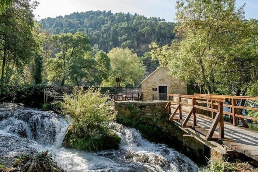 A wooden bridge over a waterfall, a stone building, and a forested hillside. 2