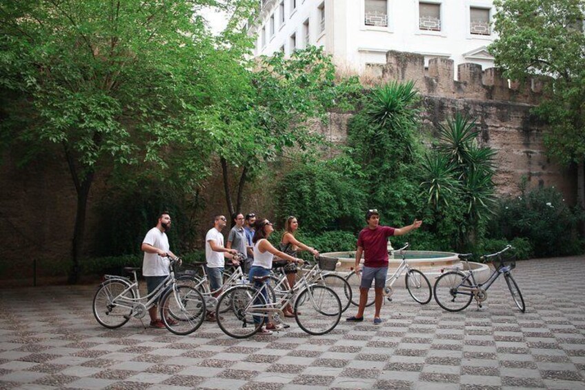 A group of people standing around bicycles in an outdoor setting with trees and a brick wall in the background. 4