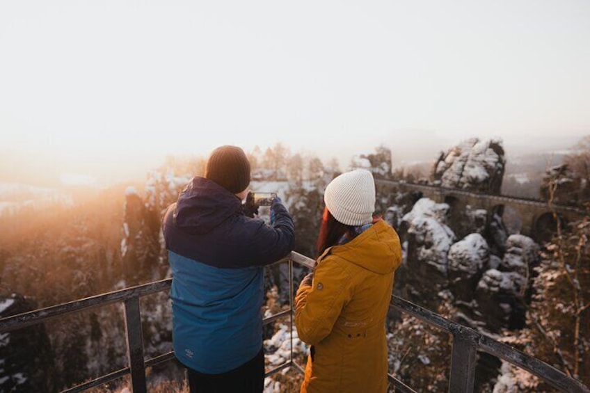 Two people taking a photo of a snowy landscape with a bridge in the background. 2