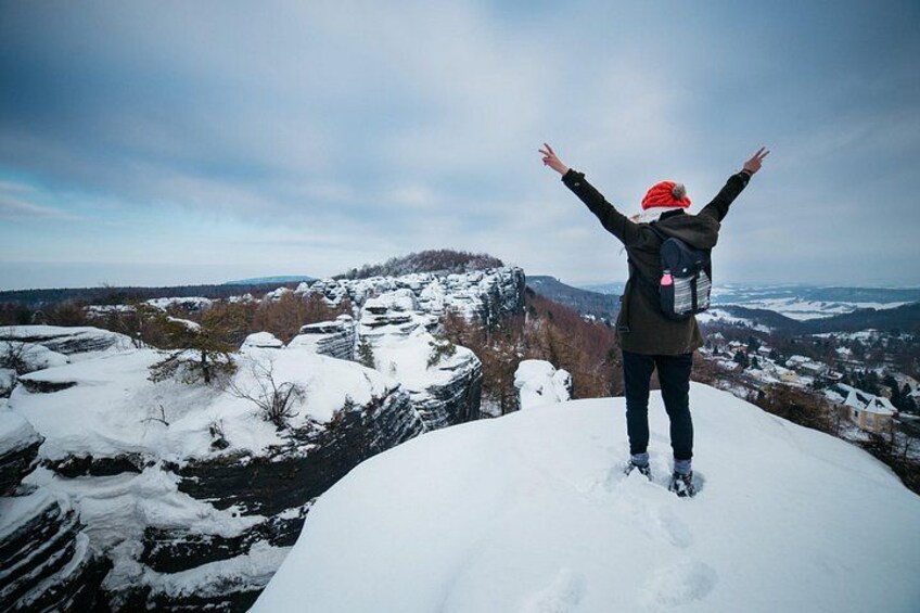 Une personne se tient debout sur un sommet enneigé, les bras levés, et surplombe un paysage hivernal parsemé d’arbres et de bâtiments. 2