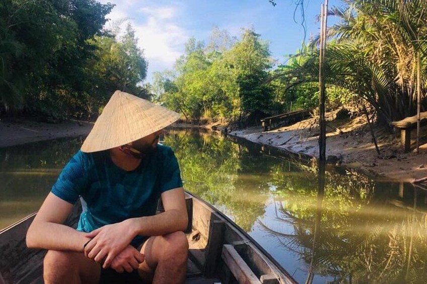 Eine Person in einem Boot, gekrönt von einem kegelförmigen Hut, umgeben von Wasser und Grünpflanzen. 3