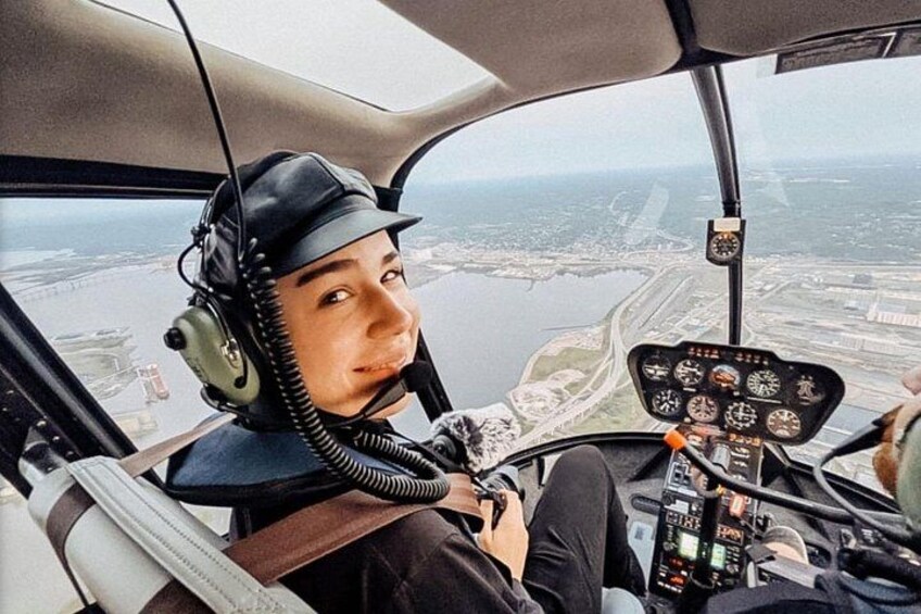 A helicopter cockpit with a pilot wearing a headset, a control panel with various gauges, and a view of a landscape with water and buildings. 1