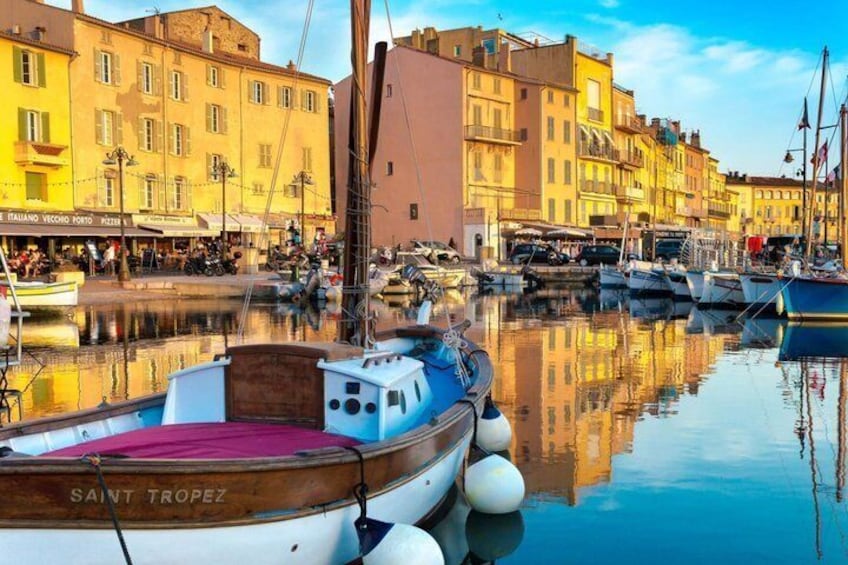 En lysthavn med fortøjede både, heriblandt en træbåd med navnet 'SAINT TROPEZ', og bygninger der spejler sig i vandet. 1