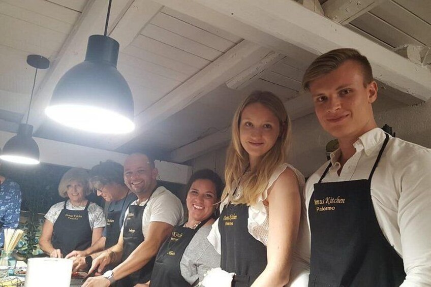 En gruppe mennesker i sorte forklæder med teksten 'Viva Kitchen Palermo', står i et køkken med hængende lamper. 1