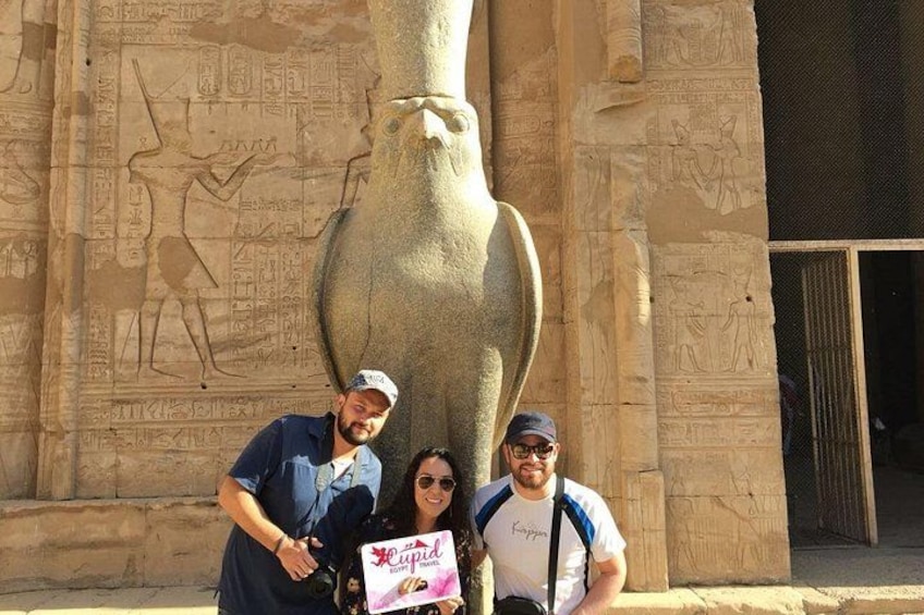 Tre persone in piedi di fronte a un'antica parete egizia con geroglifici e una grande statua di pietra. 3
