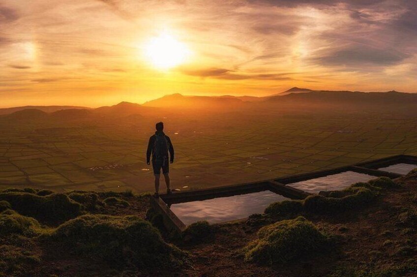 Une personne se tient debout sur une colline herbeuse, surplombant une vallée avec des champs en terrasses, et un coucher de soleil se dessine à l'arrière-plan. 1