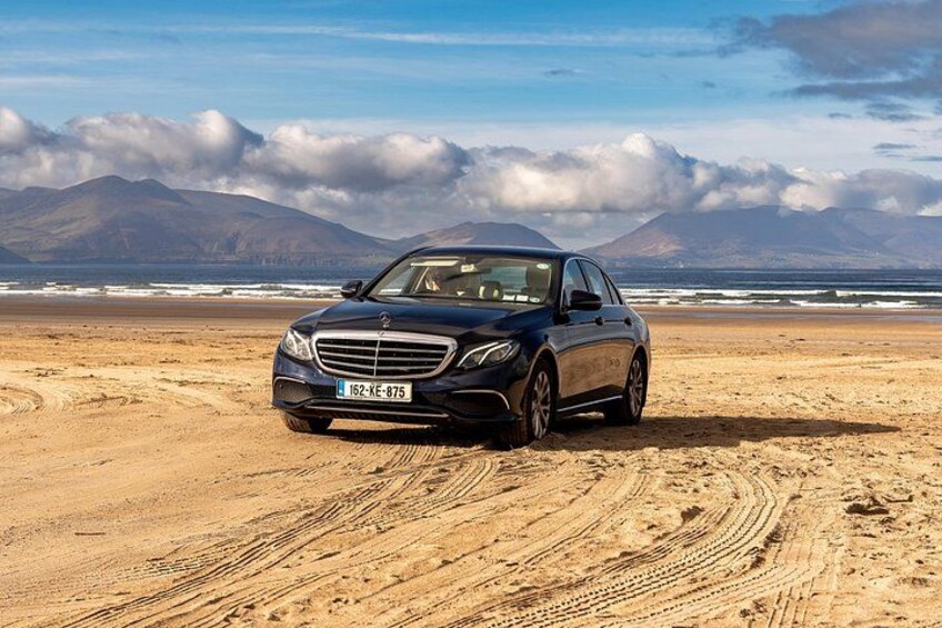 A black car parked on a sandy beach with mountains in the background. 1