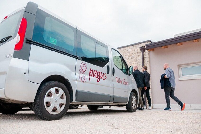 Een zilveren busje, waarop 'pagus Wine Tours' staat, geparkeerd voor een gebouw. 2