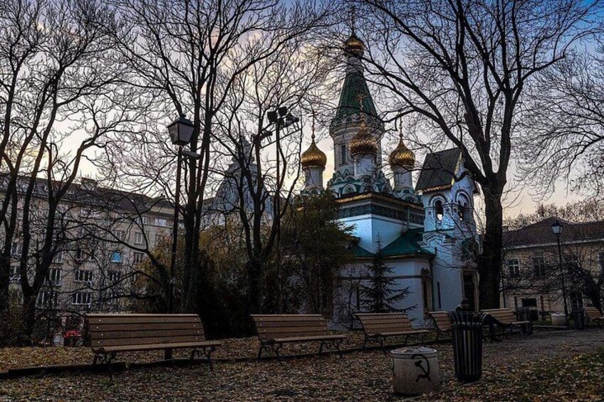 Un parque con bancos, una iglesia con cúpulas doradas y suelo cubierto de hojas. 5