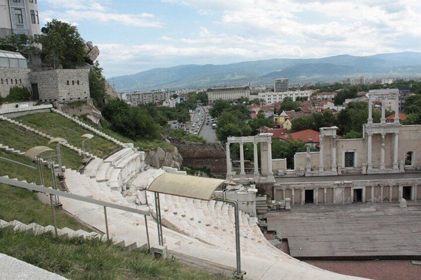 Ancient ruins with columns and a partially intact amphitheater, overlooking a cityscape with modern buildings and a distant mountain range. 5