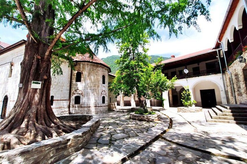 Un sendero de piedra, un árbol grande y edificios históricos con ventanas arqueadas y un tejado de baldosas. 5