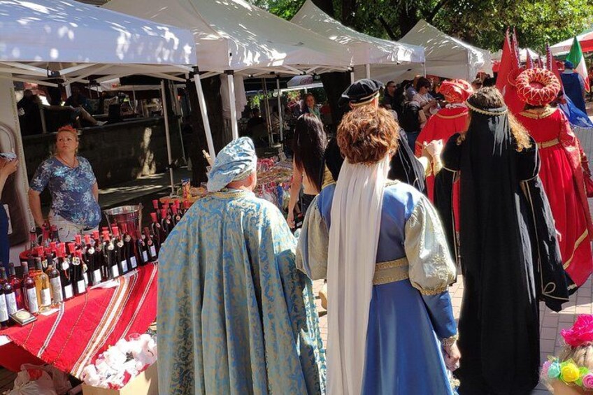 People in historical costumes at an outdoor market with wine bottles on display. 2