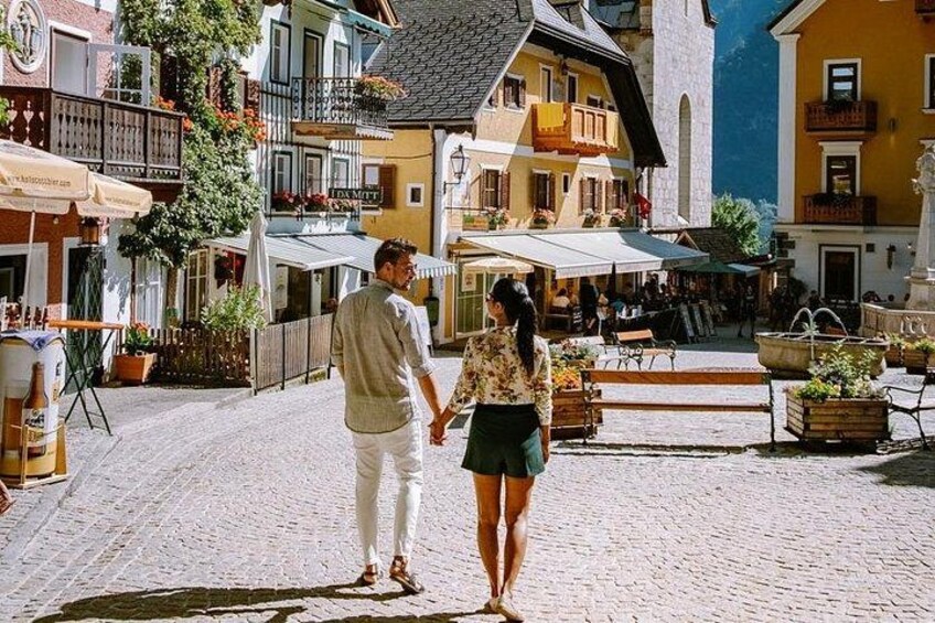 Una pareja caminando por una calle empedrada en un pueblo europeo, con edificios coloridos y terrazas al aire libre. 3