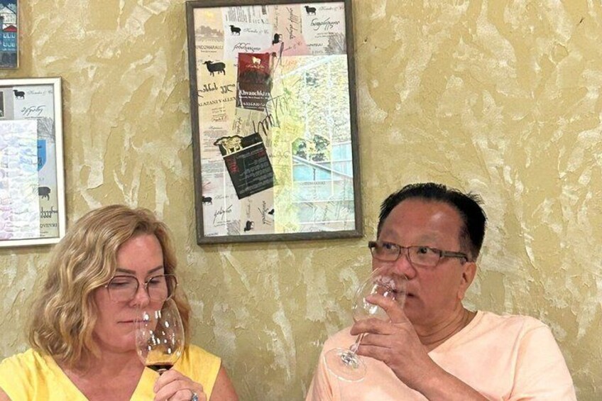 Dos personas sosteniendo copas de vino frente a un muro con un mapa y cuadros enmarcados. 1
