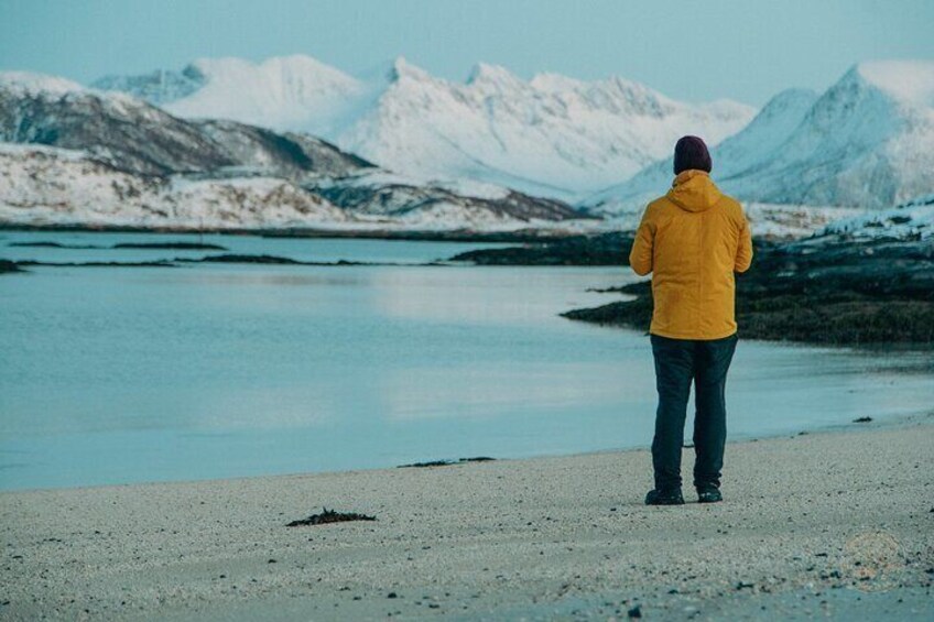 A person in a yellow jacket standing on a beach with a lake and snow-capped mountains in the background. 1