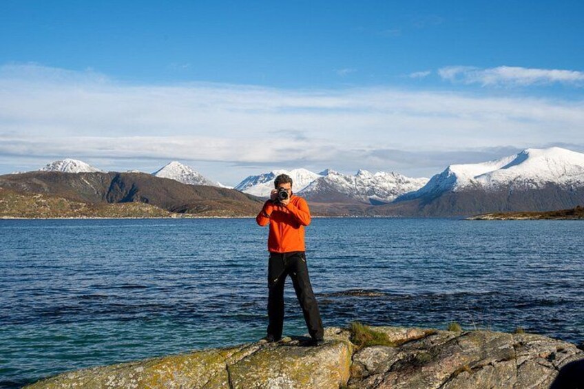 A person in an orange jacket standing on a rocky shore by a lake with snow-capped mountains in the background. 2