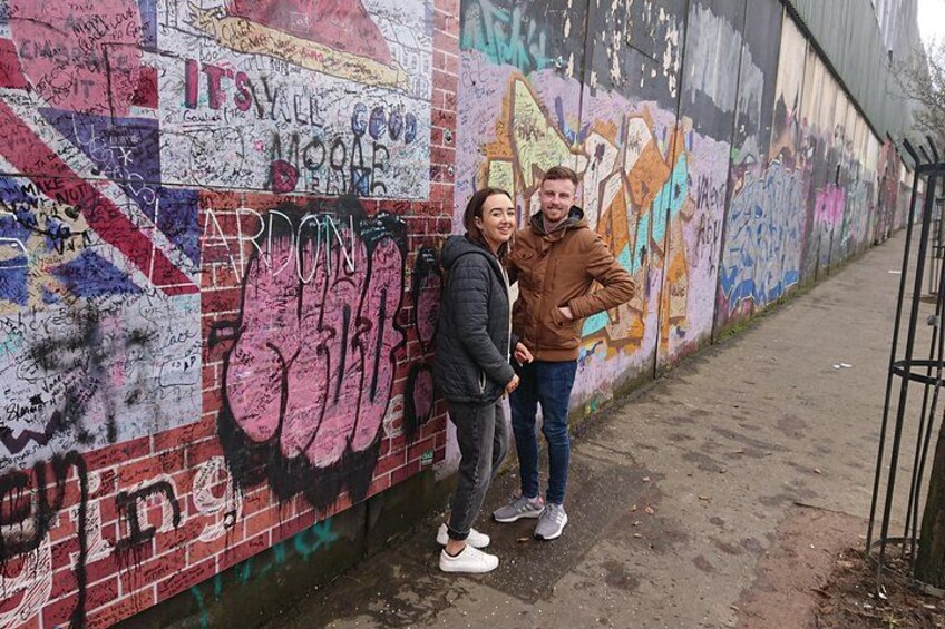 Two people standing in front of a graffiti-covered wall with various tags and messages. 3