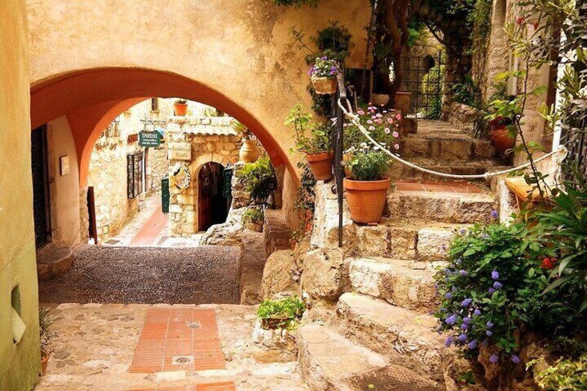 A narrow stone pathway under an archway, leading to a building entrance, with potted plants and flowers along the sides. 4