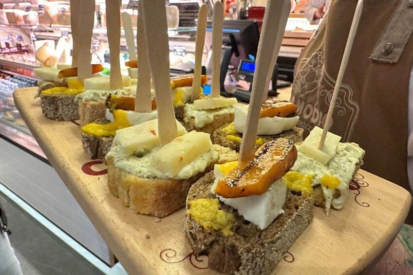 En træplade med forskellige spiser, herunder brød, ost og en grillet ret, udstillet i en butik. 1