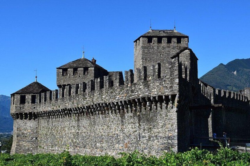 A medieval stone castle with battlements and two towers, surrounded by greenery and mountains. 2