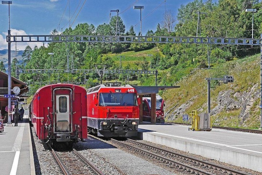 En tågstation med två röda tåg på rälsen, en perrong och ett bergigt landskap i bakgrunden. 2