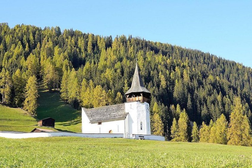 En vit kyrka med ett spetsigt torn, omgiven av ett frodigt, grönt landskap och en skogbeklädd kulle i bakgrunden. 5