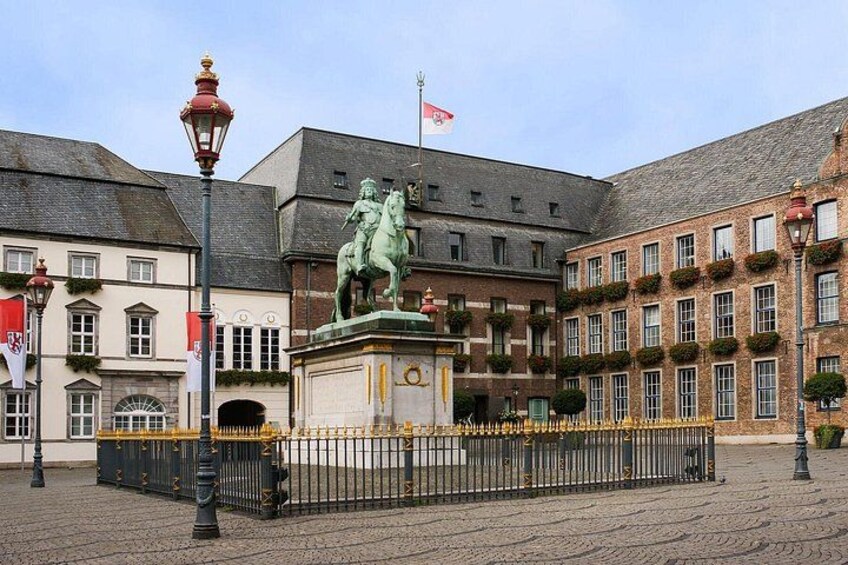 Une place historique, ornée d’une statue, bordée de bâtiments et traversée d’une rue pavée. 1