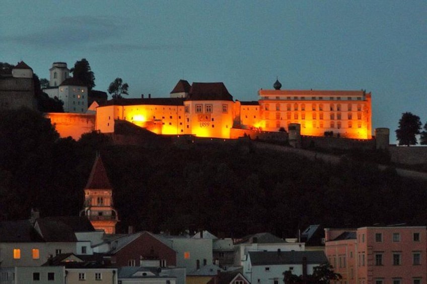 A castle with illuminated windows and a tower, surrounded by buildings and trees. 2