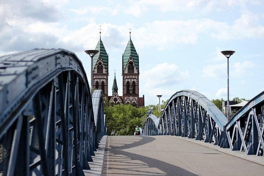A bridge with a metal structure, a church with two green spires, and a clear sky. 1