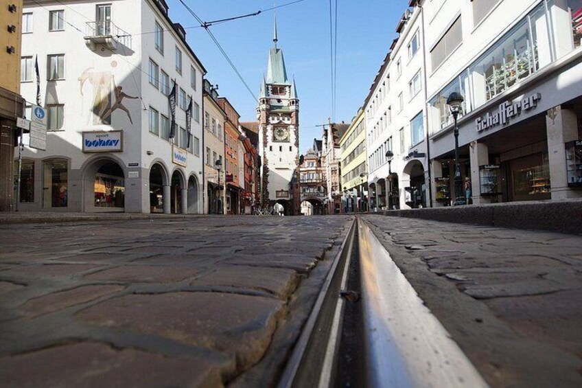 A cobblestone street with buildings, a clock tower, and a tram track. 4
