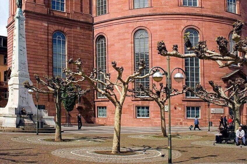 A red brick building with arched windows, a white monument, and bare trees in a plaza. 5
