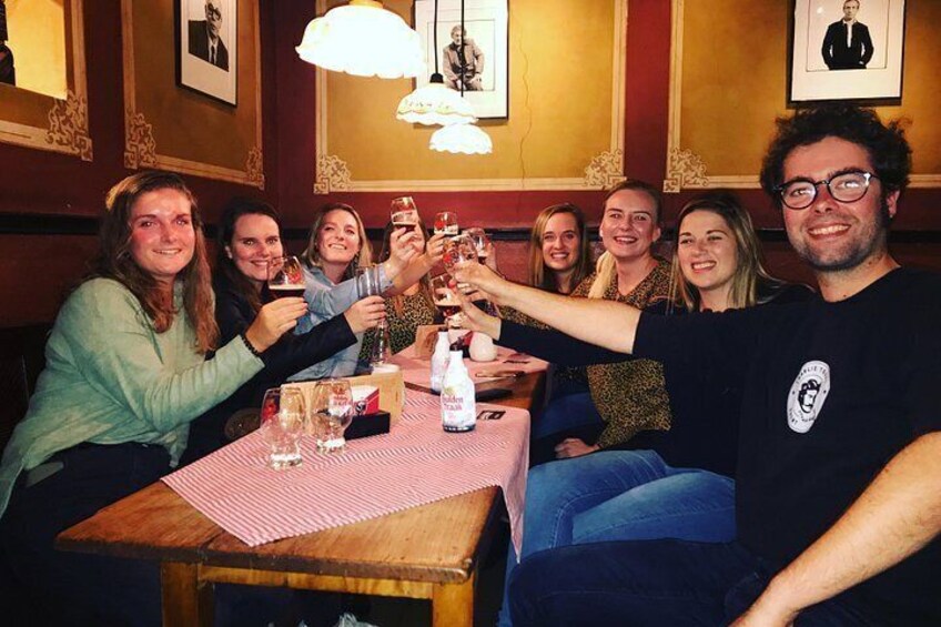 Een groep mensen proost met drankjes aan een tafel in een sfeervol verlichte kamer met ingelijste foto's aan de muur. 2