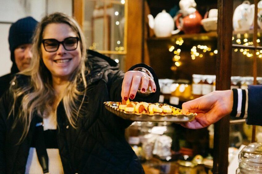 A person holding a tray with food items, another person receiving it, and a background with shelves of jars and decorative lights. 2