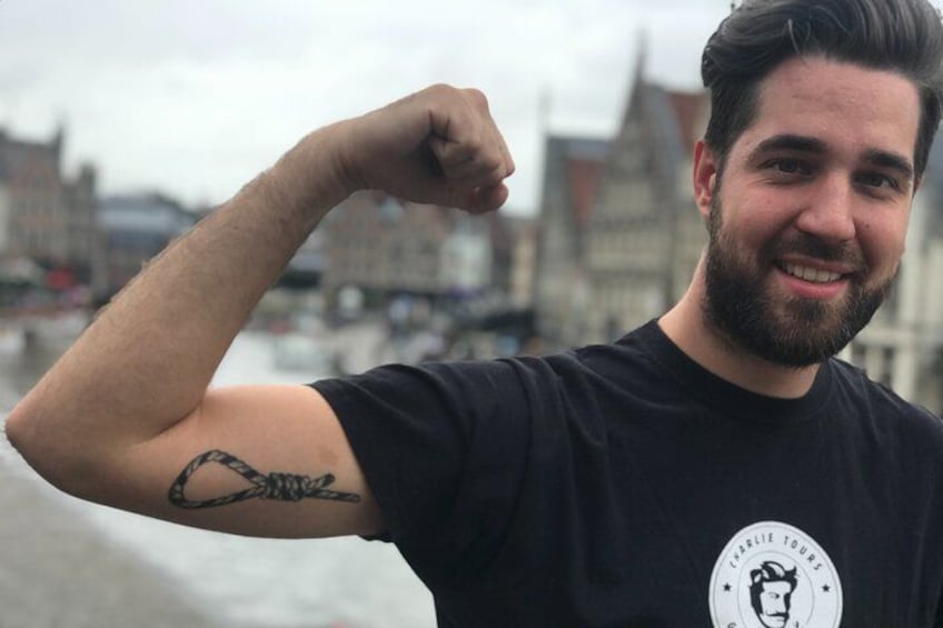En person bøjer armen med en tatovering, iført en sort t-shirt med et logo, og en byudsig i baggrunden. 5