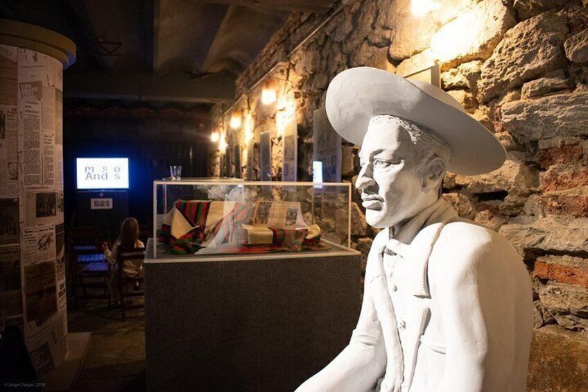 A statue of a man in a hat and suit, a stone wall, and a display case with papers and a blue screen in the background. 2