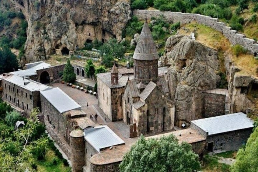 A historic stone building complex with a prominent tower, surrounded by rocky terrain and greenery. 4